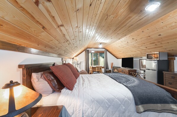 A bedroom with a wooden ceiling, a bed with pillows, a table with a lamp, a desk, a chair, a window, and a refrigerator.