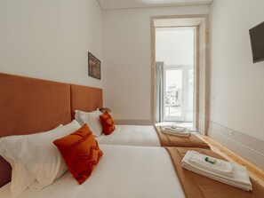 Habitació d'hotel amb dos llits, un panell de fusta al capçal, roba de llit blanca i vistes per una porta oberta.