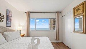 Een hotelkamer met een bed, nachtkastje, lamp, raam met gordijnen en uitzicht op zee.