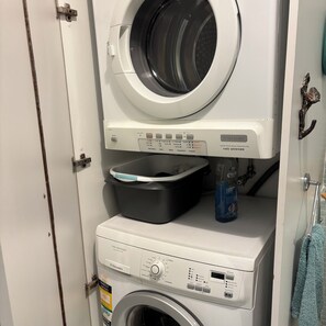 Eine Waschmaschine und ein Trockner in einem Gerät mit Bedienfeld, einer Waschmittelflasche und einem grauen Wäschekorb.