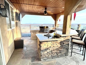 Uma cozinha externa com ilha de pedra, banquetas e vista para a paisagem.