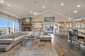 Ampla sala de estar com um sofá grande, uma mesa de centro e uma lareira. Há uma área de jantar com uma mesa e cadeiras, e uma cozinha com bancada e cadeiras.