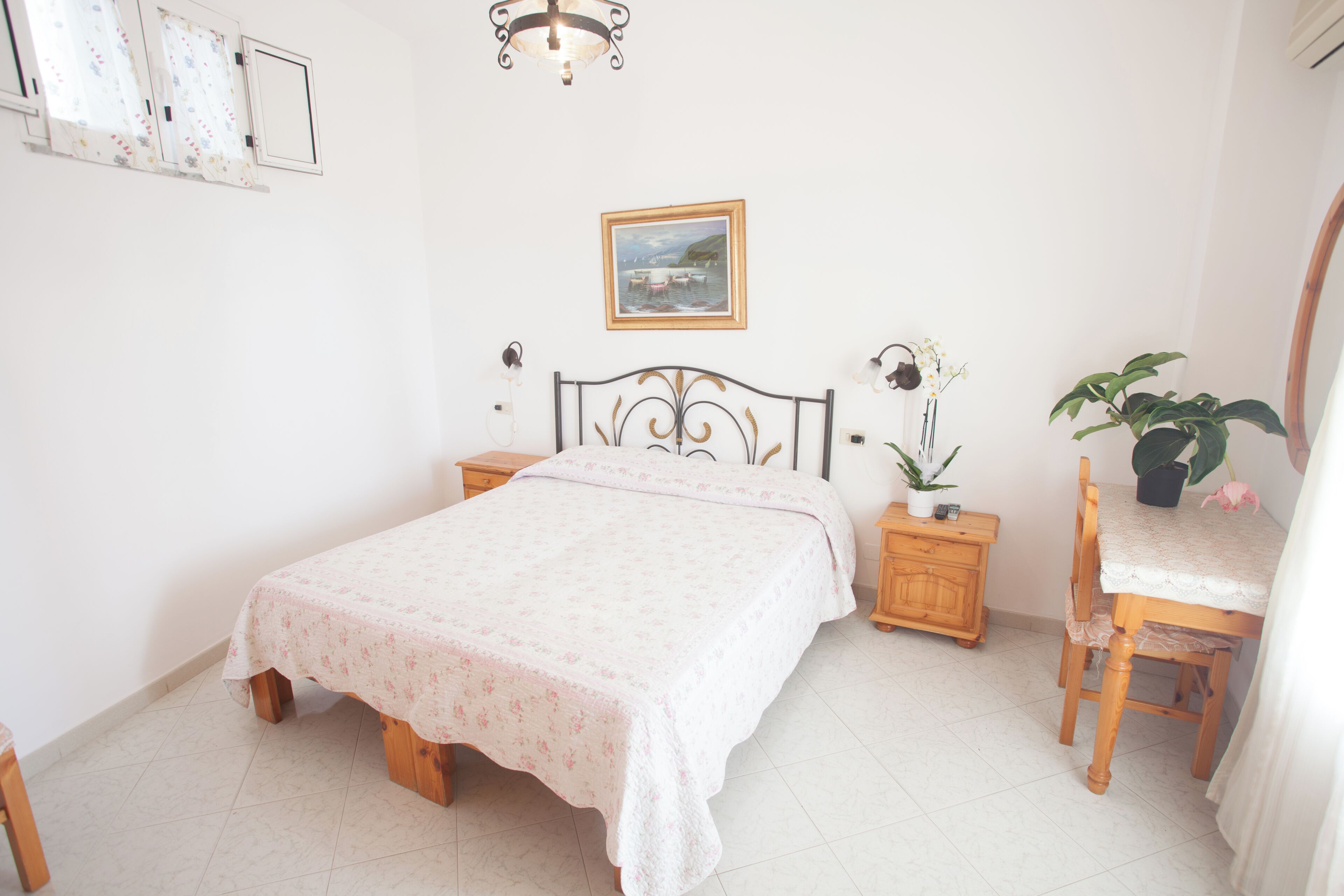 A bedroom with a bed, bedside tables, a desk with a chair, a potted plant, and a wall-mounted picture.