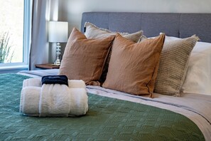 Ein ordentlich bezogenes Bett mit einer grünen Steude, mehreren Kissen und einem aufgerollten Handtuch.