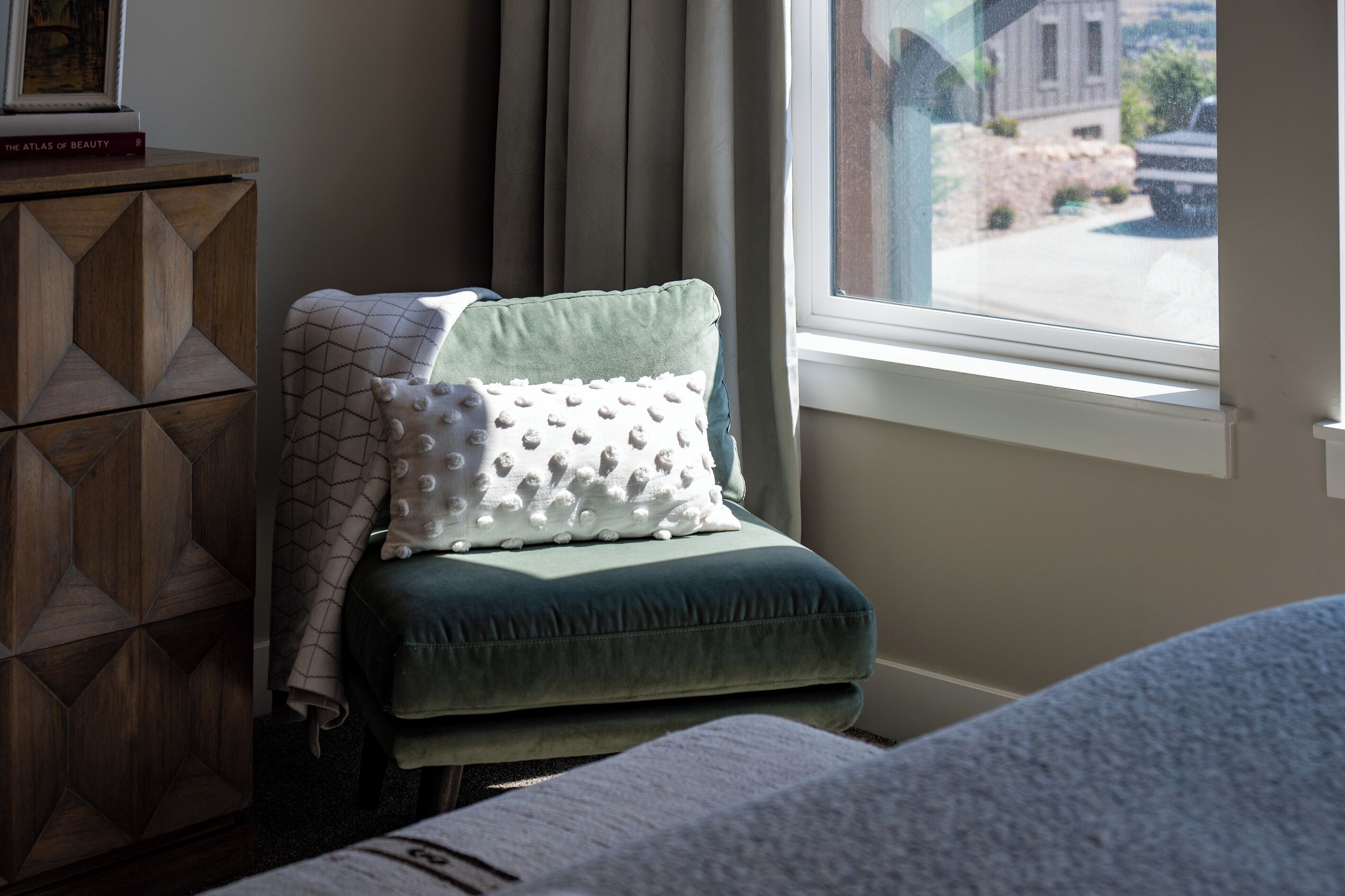 A room with a green upholstered chair, a wooden cabinet, and a window with a view of a street.