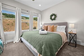 A bedroom with a large window, a bed with a green and beige bedspread, a bedside table with a lamp, and a wreath on the wall. - Eden's Basecamp |Hot Tub |Firepit | Kayaks & Bikes (Eden)