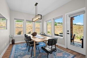 A dining area with a wooden table, black chairs, a rug, and large windows with a view of a natural landscape. - Eden's Basecamp |Hot Tub |Firepit | Kayaks & Bikes (Eden)