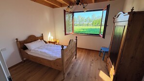 Ein Schlafzimmer mit einem hölzernen Bett, einem Nachttisch mit Lampe, einem Fenster mit Blick auf ein grünes Feld und einem blauen Sessel.