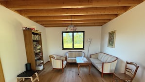 Ein Zimmer mit Holzbalkendecke, einem Bücherregal, einer Couch, einem Tisch und einem Sessel.