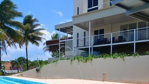 Un edificio moderno con piscina, balcón con sillas y palmeras al fondo.