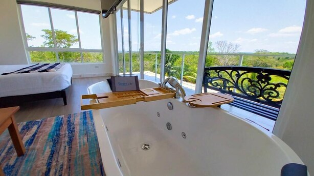 Una habitación moderna con una bañera grande, una cama y un balcón con vistas a la vegetación.