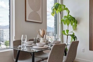 A dining area with a table set for four, a large window with a view of buildings and mountains, and a potted plant. - Stylish 2-Bedroom, 2-Bath Salt Lake City Apartment with Balcony (Salt Lake City)