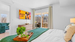 Un dormitorio con una cama, un plaid verde, una bandeja con una planta, un cuadro y vistas al exterior.