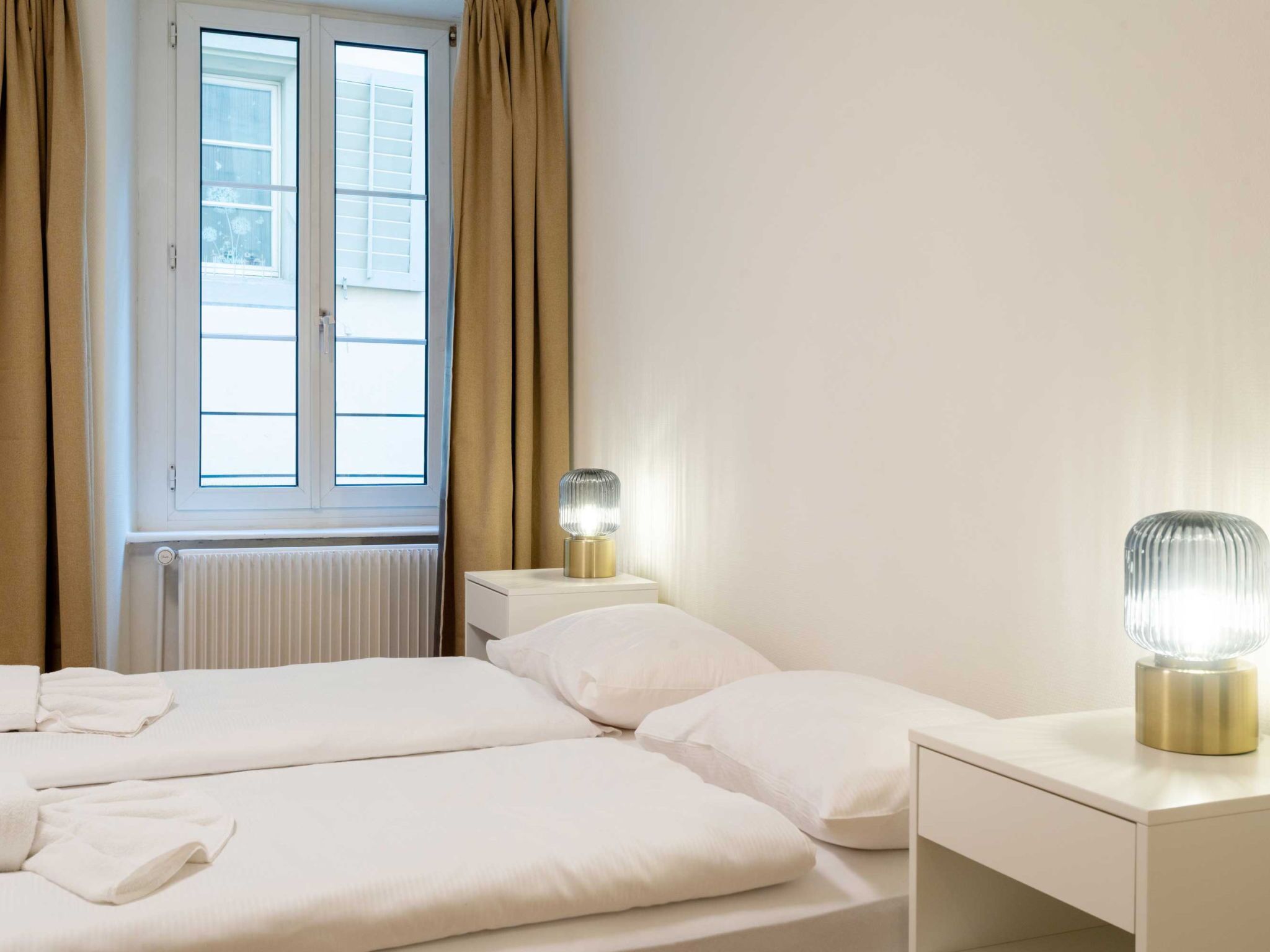 Ein Hotelzimmer mit zwei Betten, einem Fenster mit Vorhängen, einem Nachttisch mit Lampe und einer Heizung.