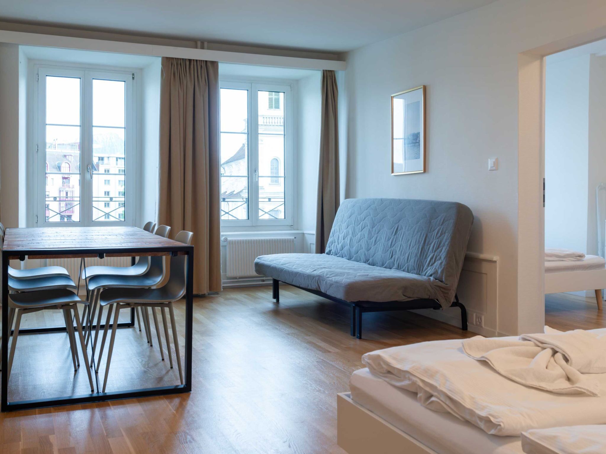 Una habitación de hotel con cama, sofá, mesa de comedor con sillas y ventanas amplias con cortinas.