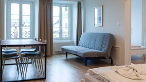 Una habitación de hotel con cama, sofá, mesa de comedor con sillas y ventanas amplias con cortinas.