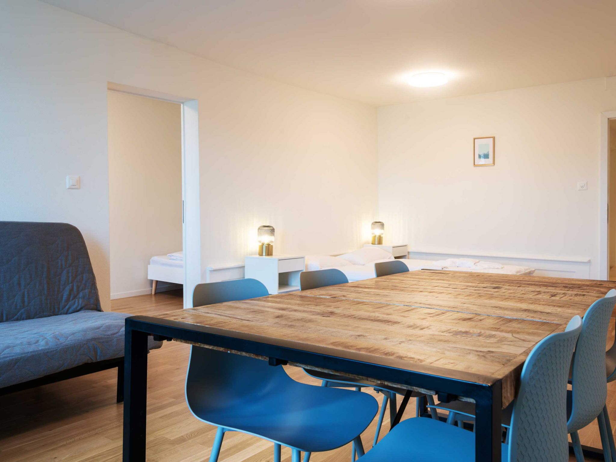 Una habitación moderna con una mesa de comedor de madera, sillas azules, un sofá gris y una cama blanca.