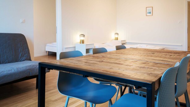 Una habitación moderna con una mesa de comedor de madera, sillas azules, un sofá gris y una cama blanca.