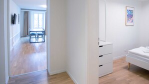 Habitación de hotel con piso de madera, una cama, una mesita de noche y un armario. Se ve una puerta que da a un comedor con una mesa y sillas.