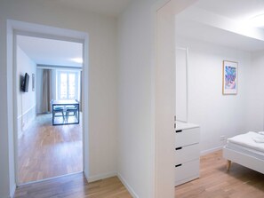 Une chambre d’hôtel avec un plancher en bois, un lit, une table de chevet et une armoire. Une porte donne sur un coin repas équipé d’une table et de chaises.