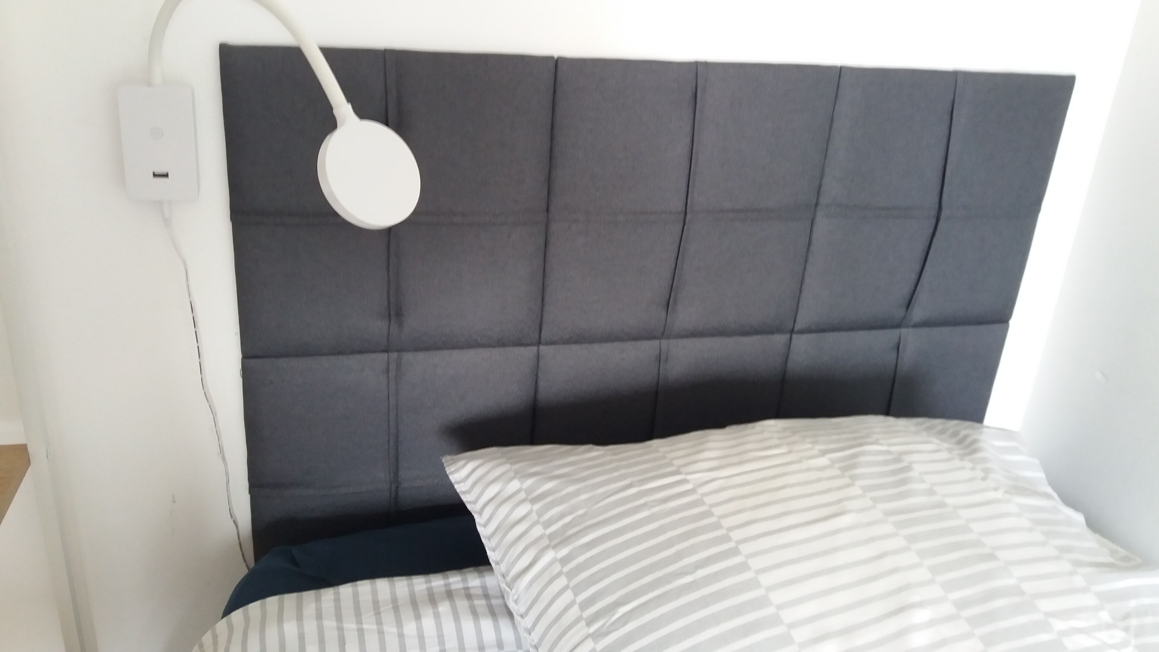 Un lit avec une tête de lit rembourrée de couleur grise, une lampe de chevet blanche et un oreiller à rayures.