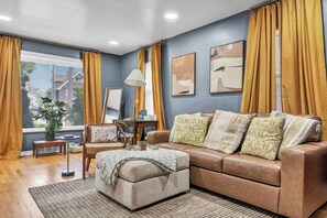 A living room with a sofa, ottoman, and a lamp. There is a window with curtains, a plant, and artwork on the wall.