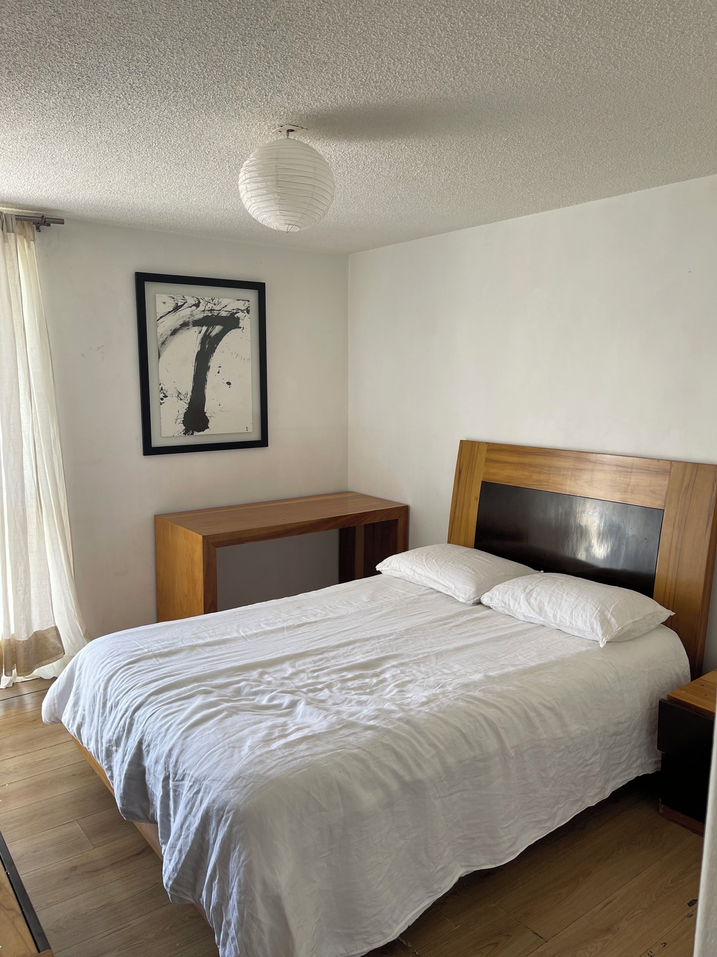 A bedroom with a bed, wooden nightstands, a white headboard, a framed artwork on the wall, and a window with curtains.