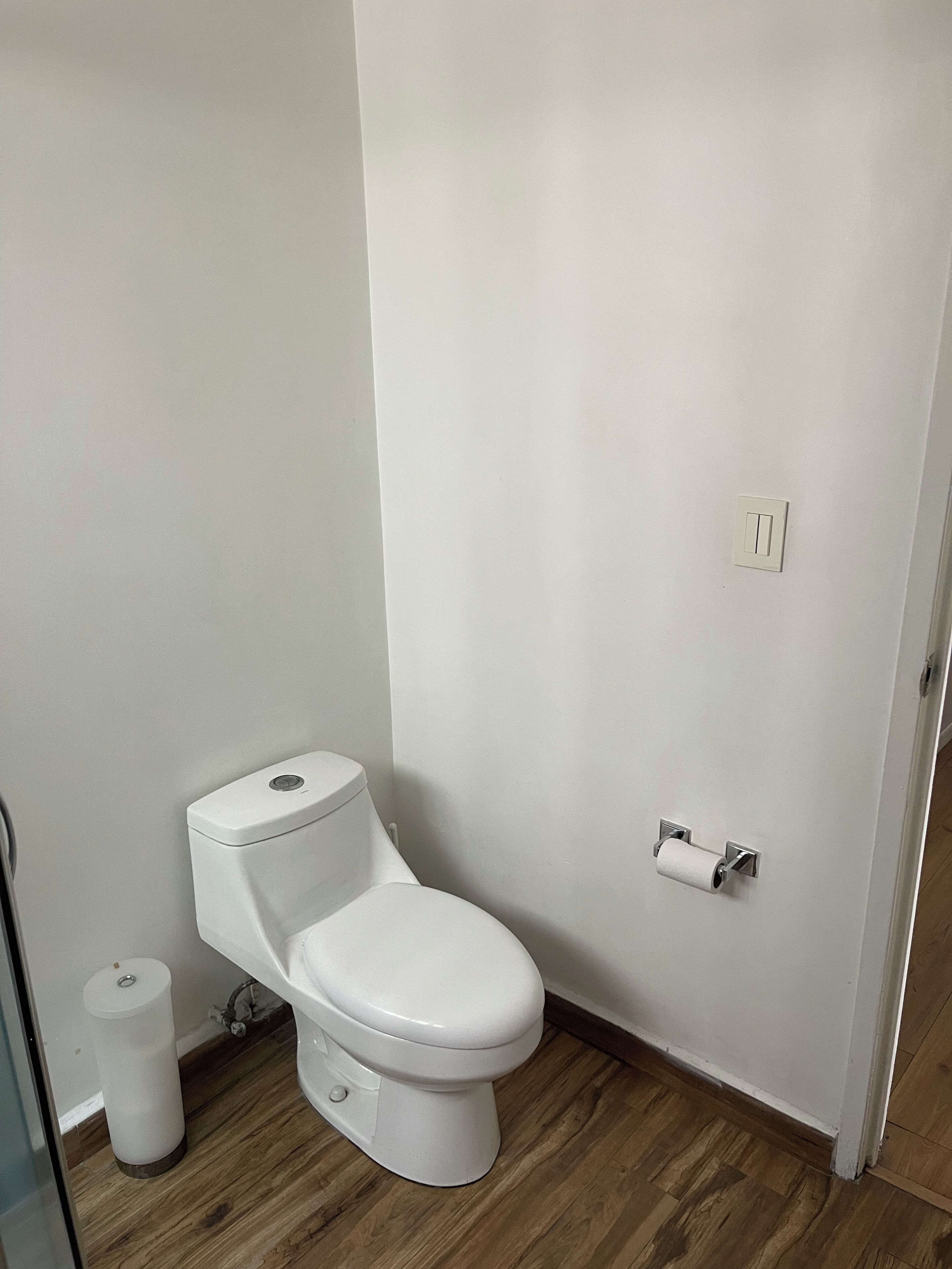 A white toilet with a flush handle, a toilet paper holder, and a small trash bin.