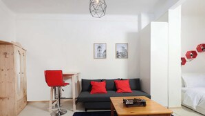 Een kamer met een donkergrijze bank, rode kussens, een houten tafel, een rode stoel, een witte kledingkast en twee ingelijste foto’s aan de muur.