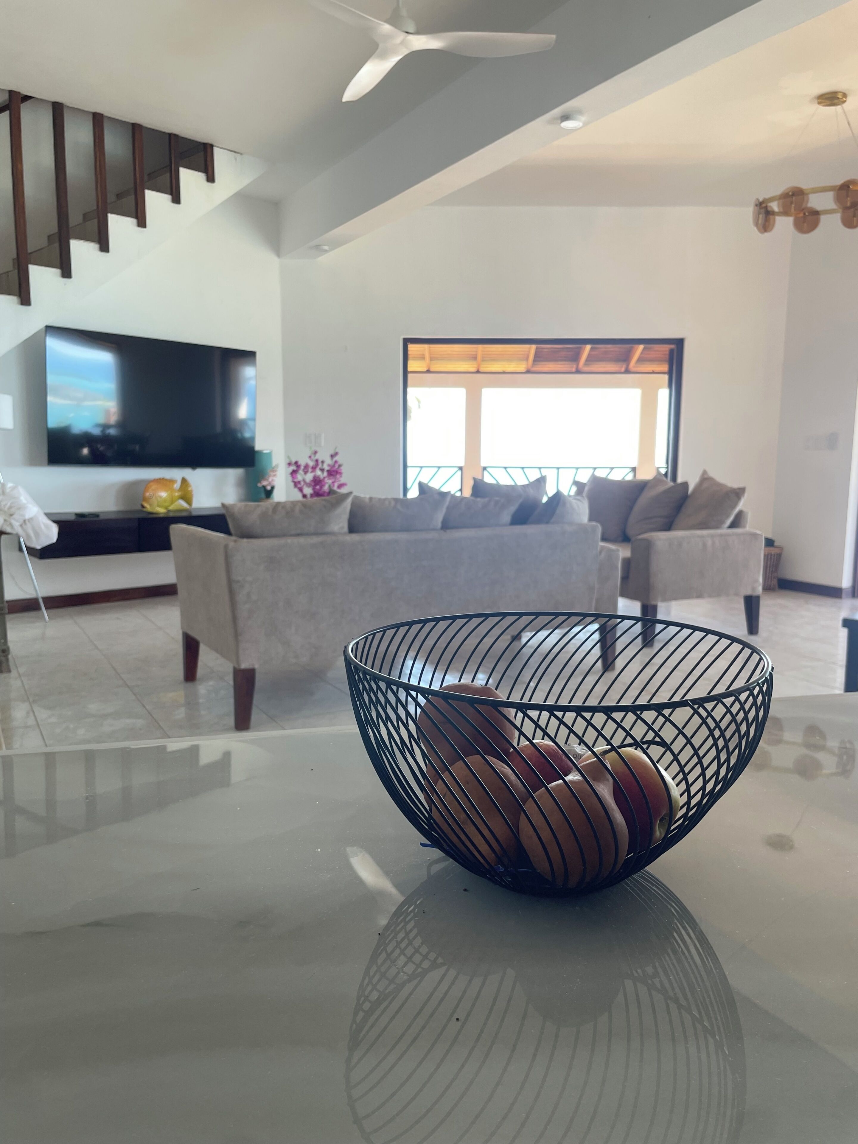 A modern living room with a flat-screen TV, a sofa, a coffee table with a wire bowl, and a staircase.