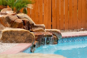 A pool with a rock waterfall feature and a wooden fence in the background. - Shady Oak: Pool, Hot Tub, Game Room | Sleeps 12 (Live Oak)