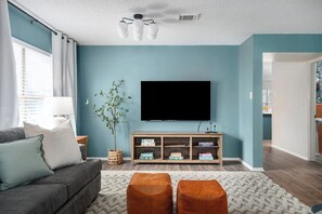 A living room with a TV, sofa, ottomans, and a bookshelf. - Shady Oak: Pool, Hot Tub, Game Room | Sleeps 12 (Live Oak)