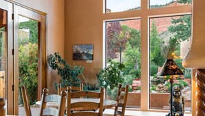 A dining area with wooden chairs and a round table, large windows with a view of greenery, and a ceiling with exposed beams.