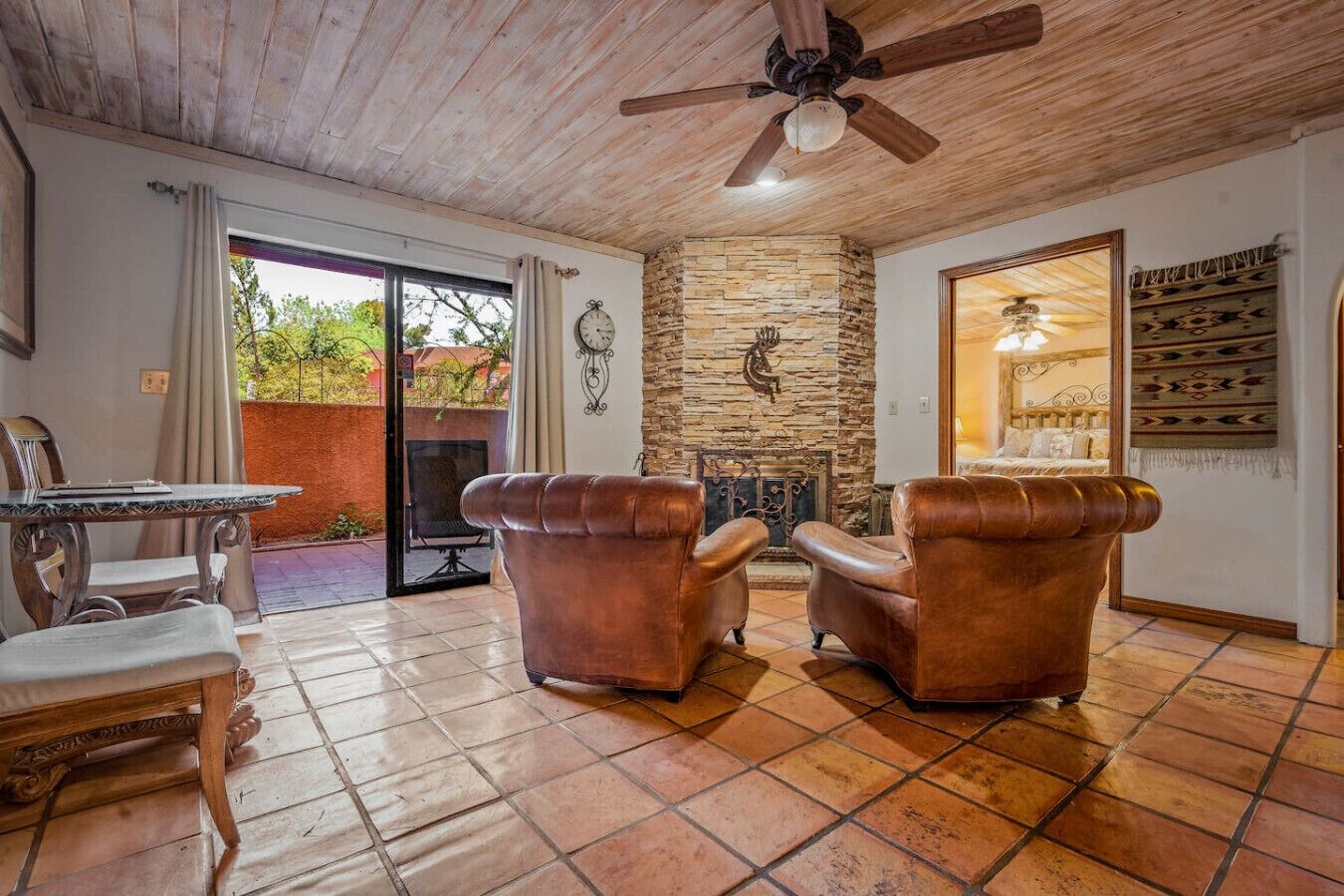 A room with a stone fireplace, leather armchairs, a dining table with chairs, and a ceiling fan.