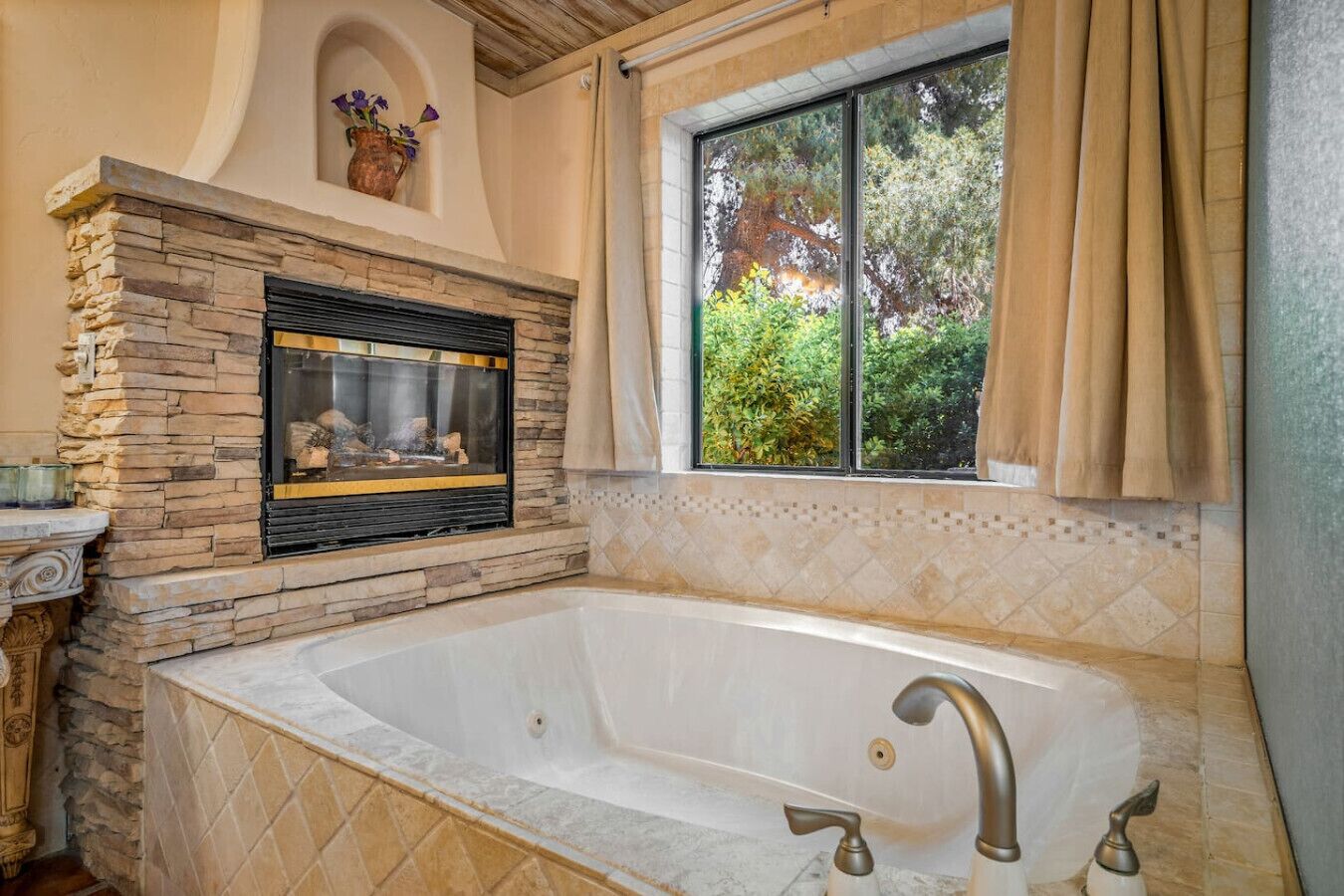 A bathroom with a stone fireplace, a large bathtub, and a window with curtains.