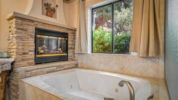 A bathroom with a stone fireplace, a large bathtub, and a window with curtains.