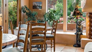 A dining area with wooden chairs and a table, large windows with a view of greenery, and a statue on a pedestal.