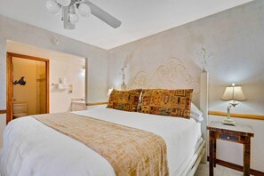 Un dormitorio con cama, mesita de noche, lámpara, ventilador de techo y una puerta que da a un baño.