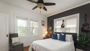 Un dormitorio con una cama, una silla, un ventilador de techo y una guitarra en la pared.
