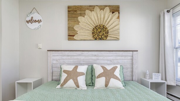 A bedroom with a bed, starfish pillows, a welcome sign, and a sunflower artwork. - King Suite Getaway, Downtown Ogunquit. Walk to Beach, Shops & Dining! (Ogunquit)