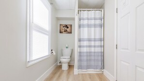 A narrow bathroom with a toilet, a window with blinds, a shower curtain, and a framed picture on the wall. - King Suite Getaway, Downtown Ogunquit. Walk to Beach, Shops & Dining! (Ogunquit)