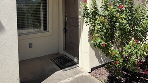 La entrada de una casa con una puerta marcada '19', un ventanal con persianas y un pequeño jardín con flores rojas.