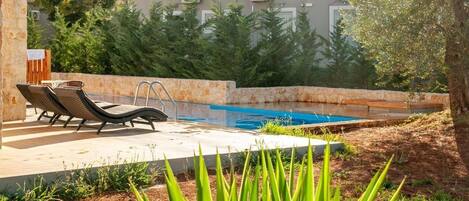 A swimming pool with lounge chairs, a concrete deck, and a building with air conditioning units in the background.