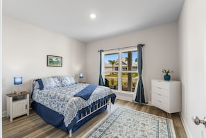 Un dormitorio con cama, mesitas de noche, cómoda, ventana con cortinas, alfombra y un cuadro en la pared.