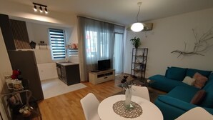 A modern living room with a teal sofa, a white dining table with chairs, a kitchen area with a refrigerator, and a TV.