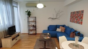 A living room with a blue sofa, a coffee table, a TV on a wooden stand, a white table with a vase, and a wall-mounted air conditioner.