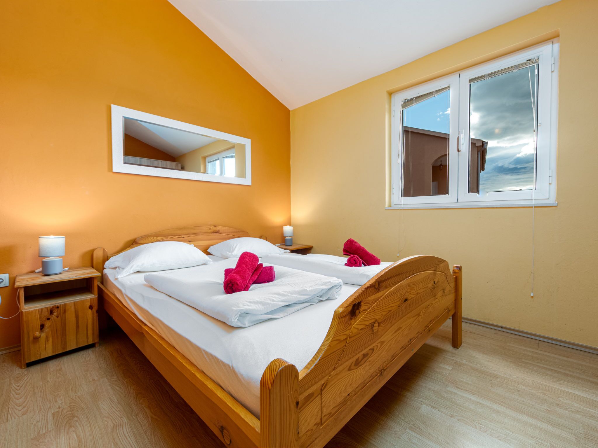 Une chambre individuelle avec un lit en bois, une table de chevet, une commode, un miroir et une fenêtre donnant sur le ciel.