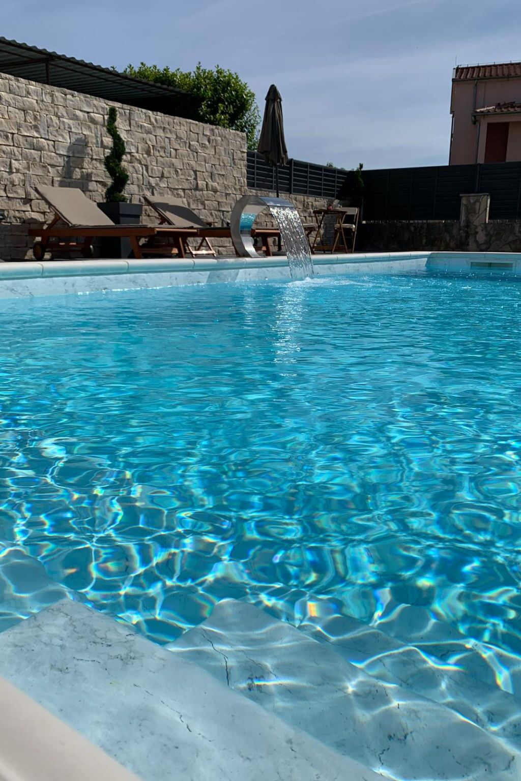 A swimming pool with clear blue water, lounge chairs, and a stone wall.
