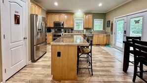 A kitchen with wooden cabinets, stainless steel appliances, a central island, and a dining area with chairs. - La Buena Vida Retreat  Hot Tub • Game Room • Fire Pit (Huddleston)