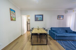A room with a dining table, chairs, a blue sofa, and two framed pictures on the wall. - Apto a 750m da Praia da Enseada com Ar e Wi-fi (Guarujá)